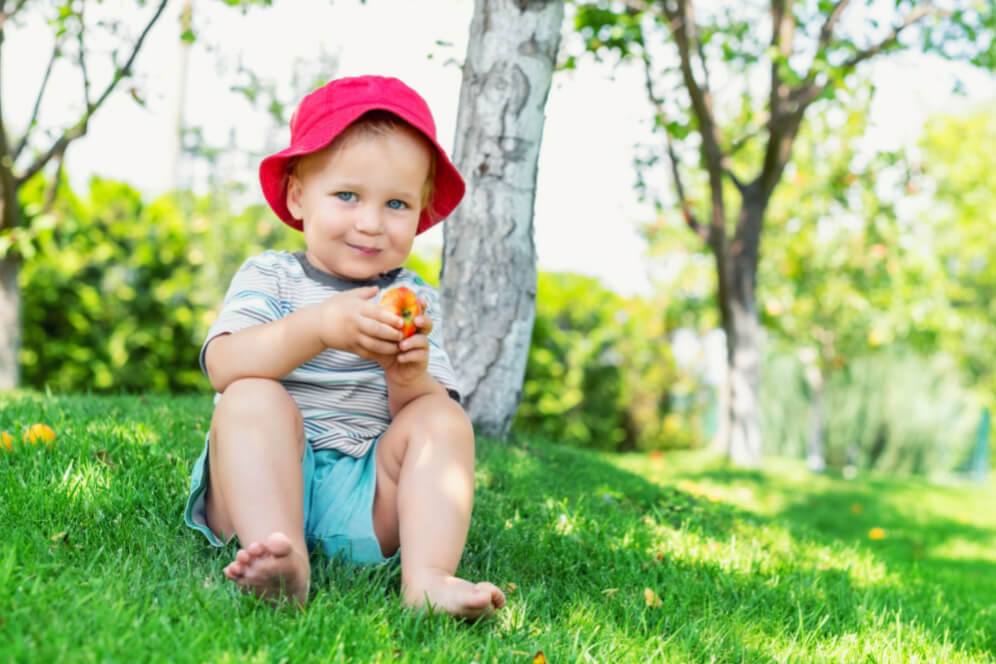 copil descult sta in sezut pe iarba in parc cu un mar in mana si palarie de protectie rosie
