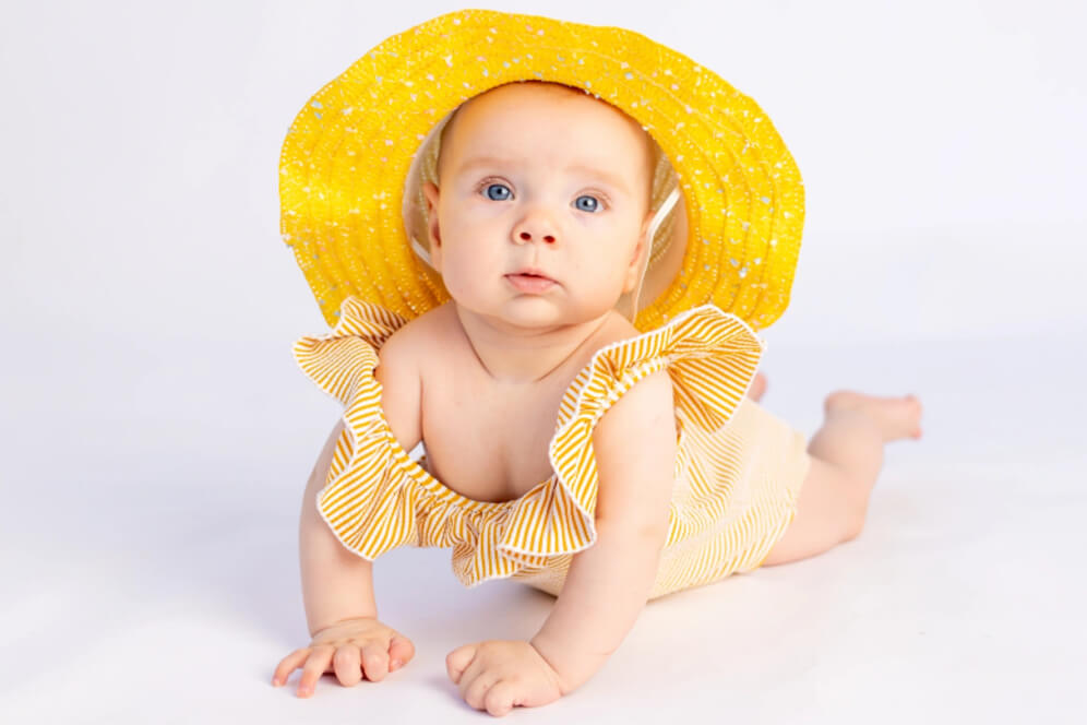 bebelus stand pe burta, imbracat in galben cu o palarie mare de soare tot galbena