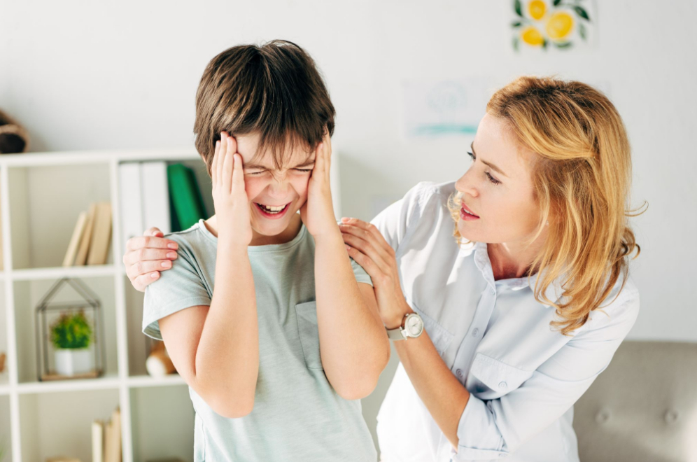 copil cu sindrum autist cu mainile la cap, tipa si terapeuta incearca sa il linisteasca