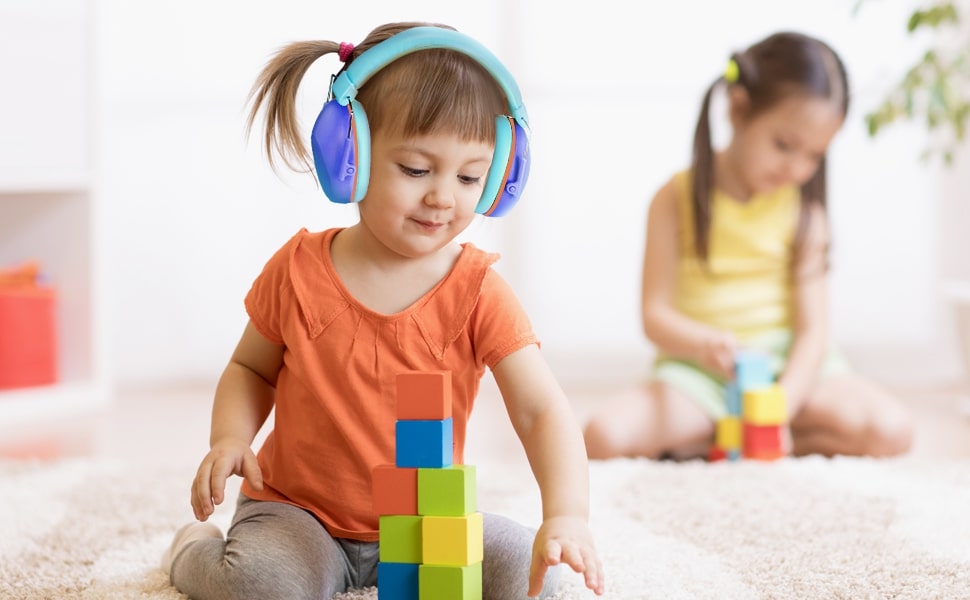 fetita cu casti antifonice in urechi construieste un turn din cuburi, in fundal mai este o alta fetita care face la fel