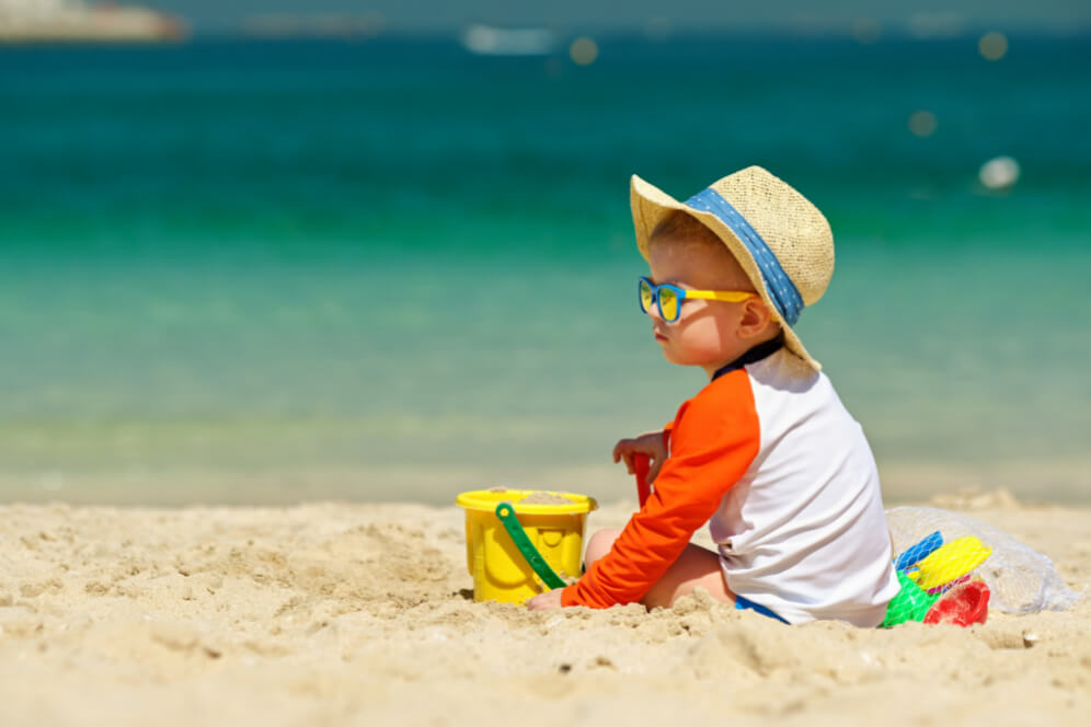copil pe plaja jucandu-se in nisip cu palarie de soare pe cap 