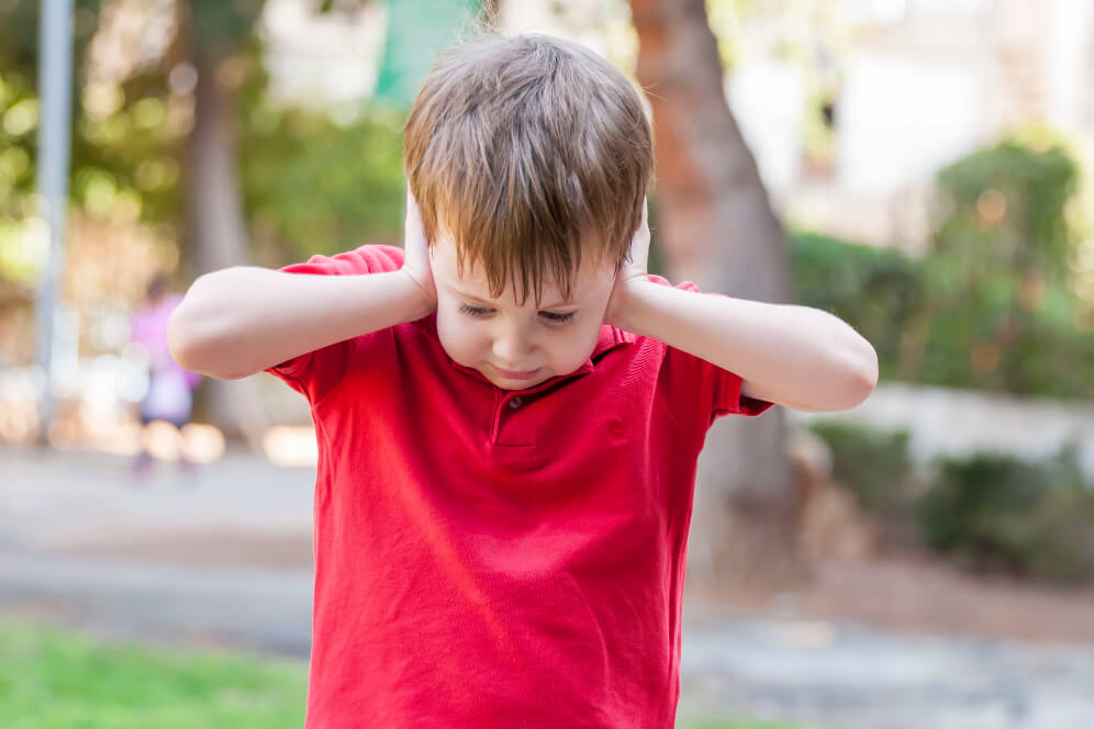 copil in tricou rosu care se afla in parc si tine mainile la urechi pentru ca este zgomot puternic