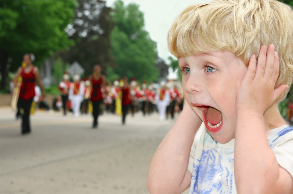 copil blond cu ochi albastri care tine mainile la urechi si tipa in timp ce in fundal este o parada zgomotoasa